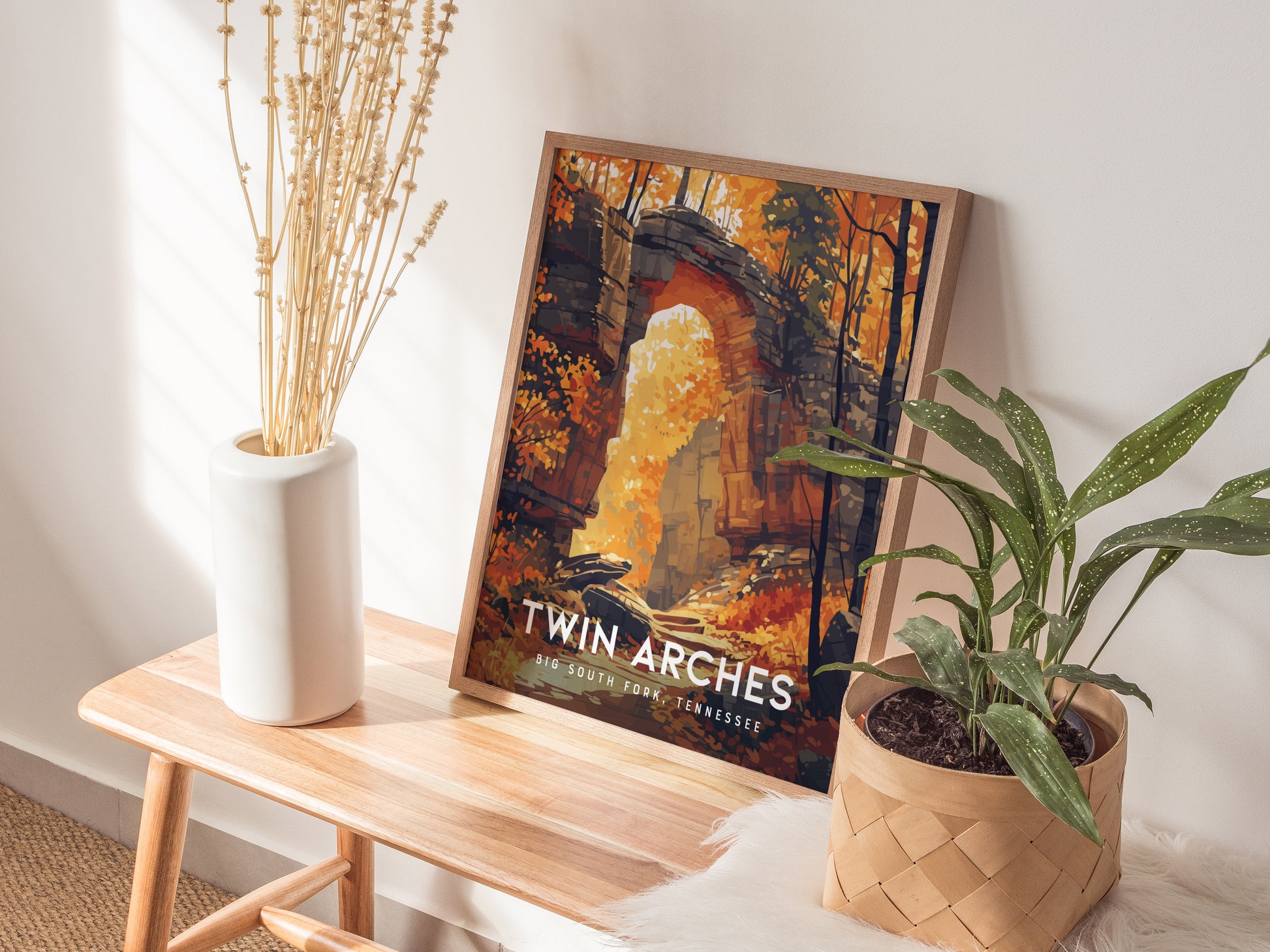A framed Twin Arches poster rests on a wooden bench beside a white vase and a potted plant, illuminated by natural light.