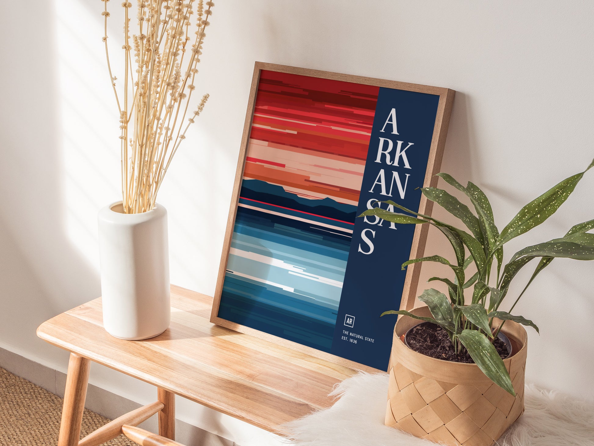 A minimalist Arkansas poster with bold, abstract landscape colors is displayed on a wooden bench, accompanied by a white vase with dried flowers and a potted green plant.