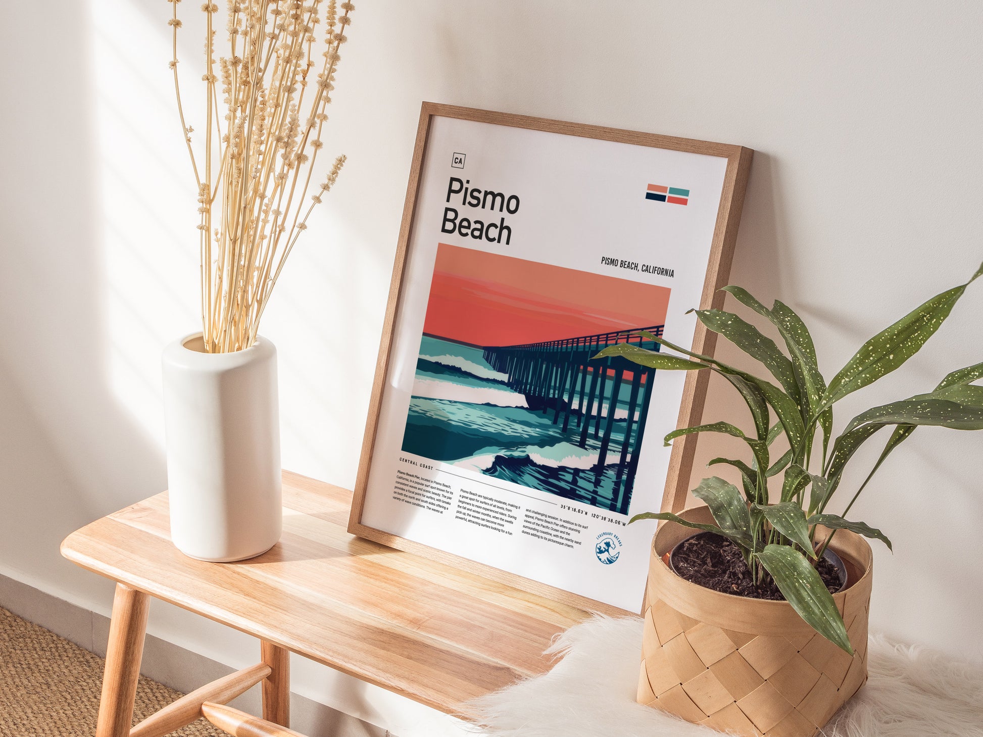 A framed Pismo Beach poster with a pier and sunset design rests on a wooden bench, accompanied by a white vase with dried flowers and a potted plant.