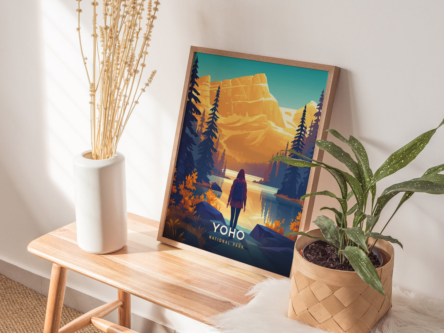 A framed Yoho National Park poster depicting a vibrant landscape with mountains and a hiker is displayed on a wooden bench, accompanied by a vase of dried flowers and a.