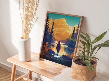 A framed Yoho National Park poster depicting a vibrant landscape with mountains and a hiker is displayed on a wooden bench, accompanied by a vase of dried flowers and a.
