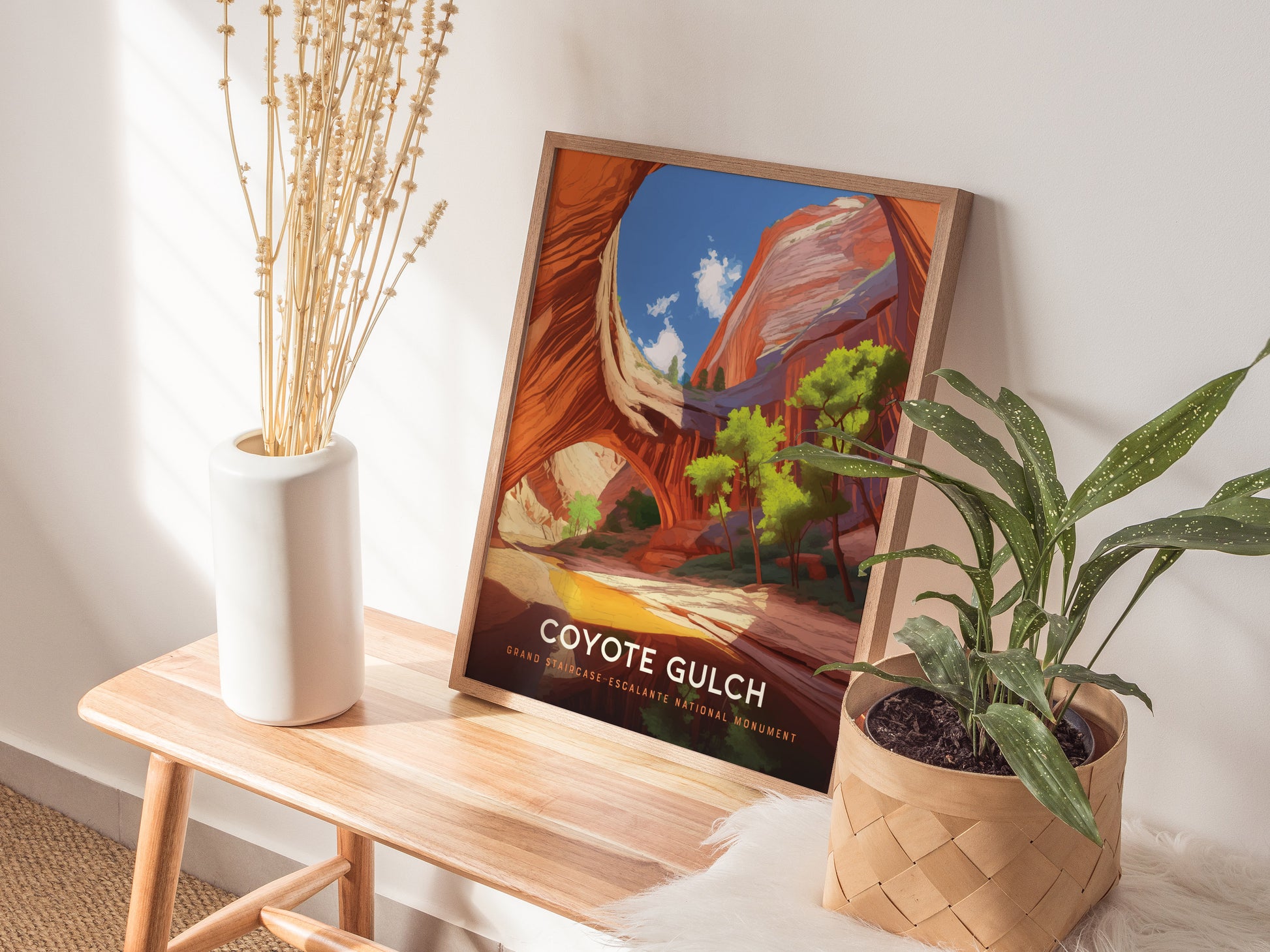 A framed Coyote Gulch poster leans against a wall on a wooden bench, surrounded by a white vase with dried flowers and a potted plant.