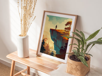 A framed Acadia National Park poster rests on a wooden bench, surrounded by a white vase with dried plants and a potted green plant. Sunlight casts soft shadows.