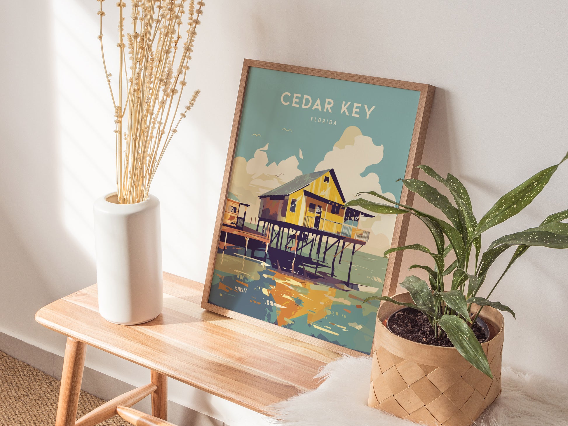 A Cedar Key, Florida poster depicting a stilt house over water is propped against a wall on a wooden bench, next to a white vase with dried flowers and a.