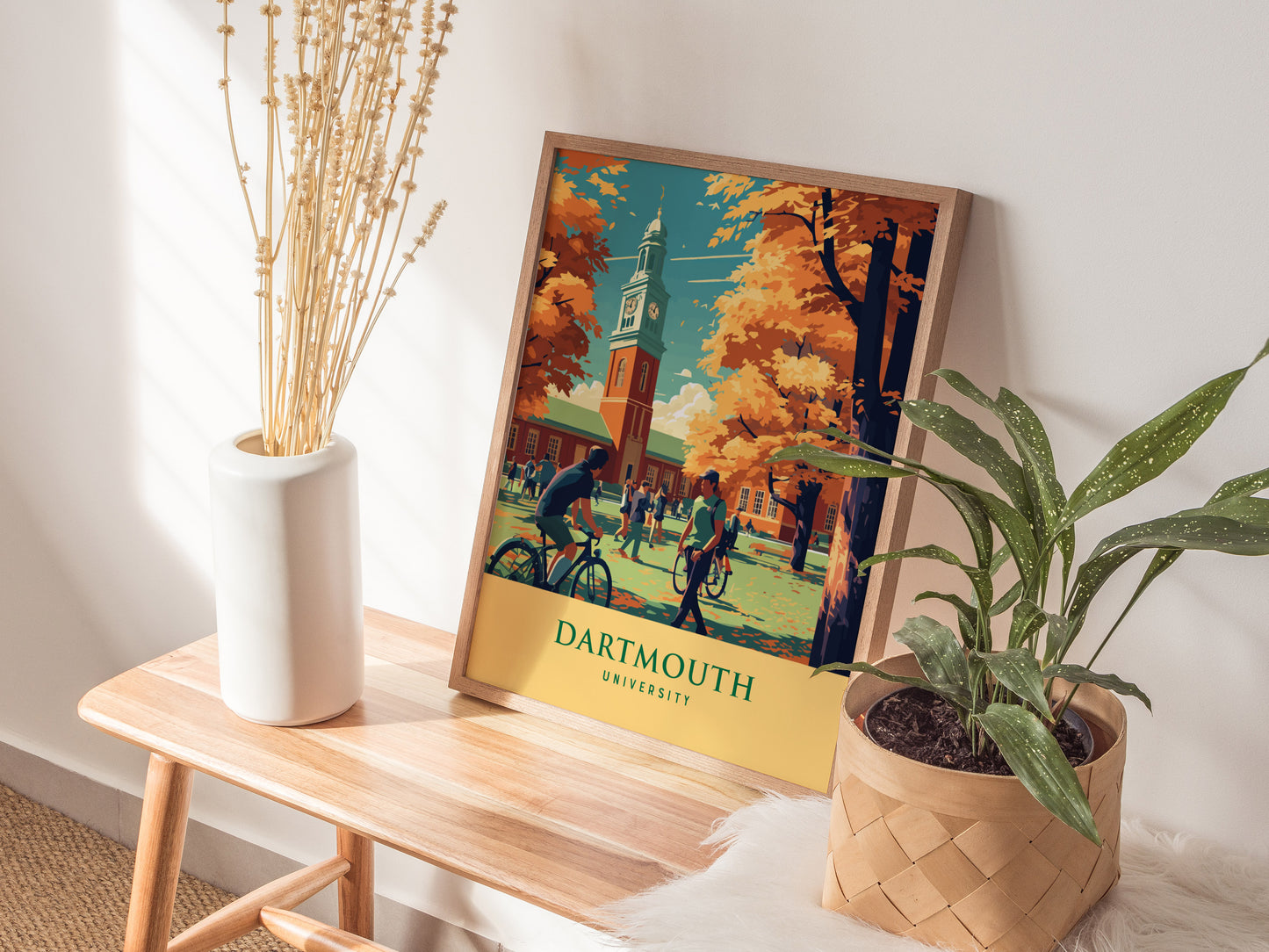 A Dartmouth University poster featuring students and autumn trees is displayed on a wooden bench, accompanied by a white vase with dried plants and a potted green plant.
