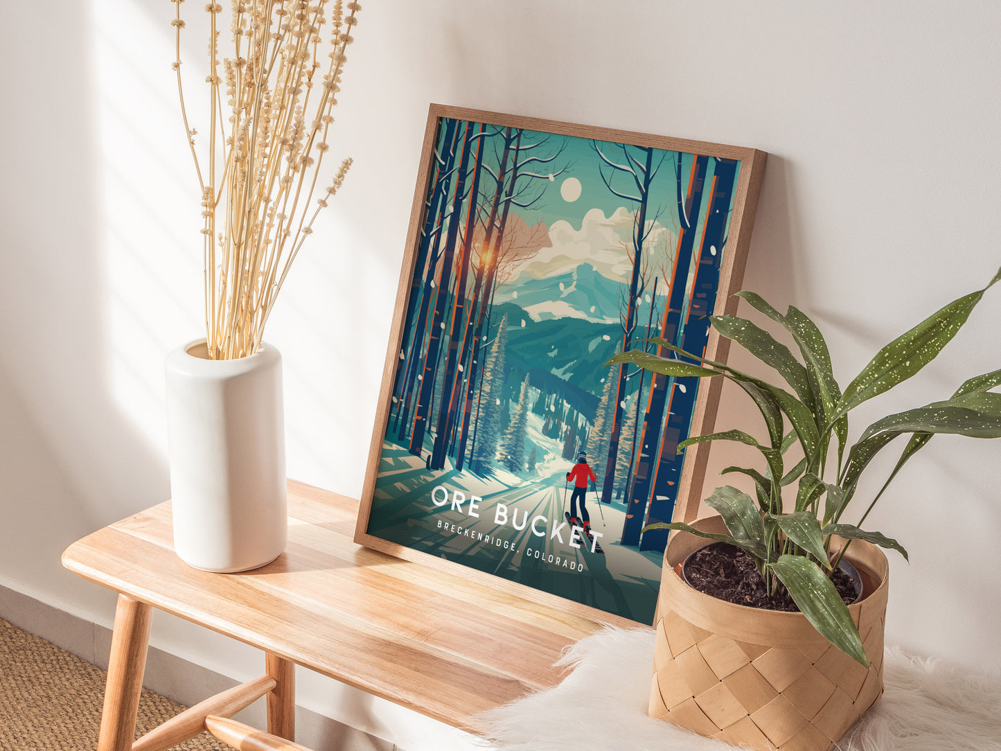 A framed Ore Bucket poster featuring a snowy mountain scene and skier rests on a wooden bench, surrounded by a white vase with dried stems and a potted green plant.