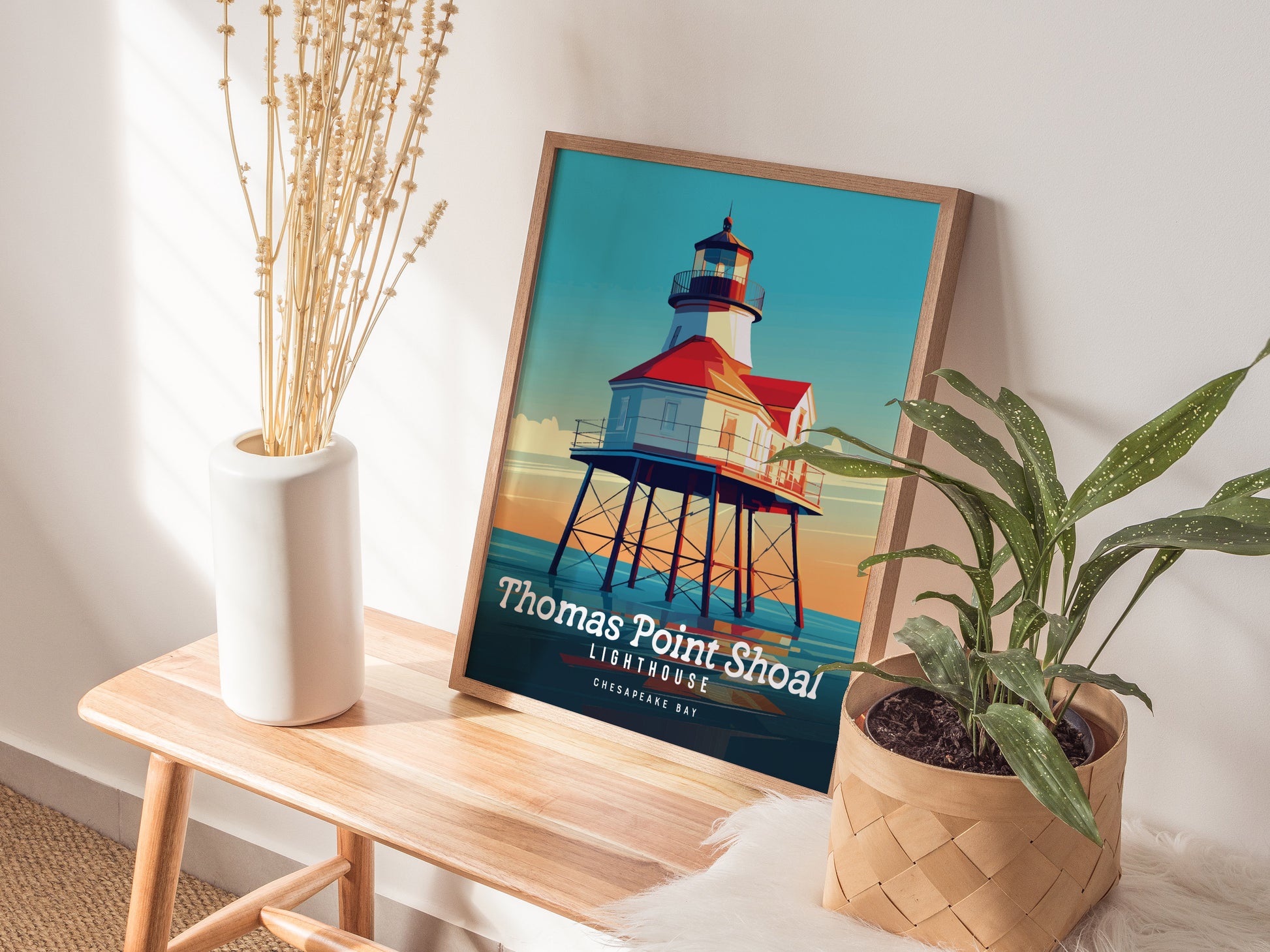 A framed Thomas Point Shoal Lighthouse poster is displayed on a wooden bench beside a white vase with dried flowers and a green potted plant.