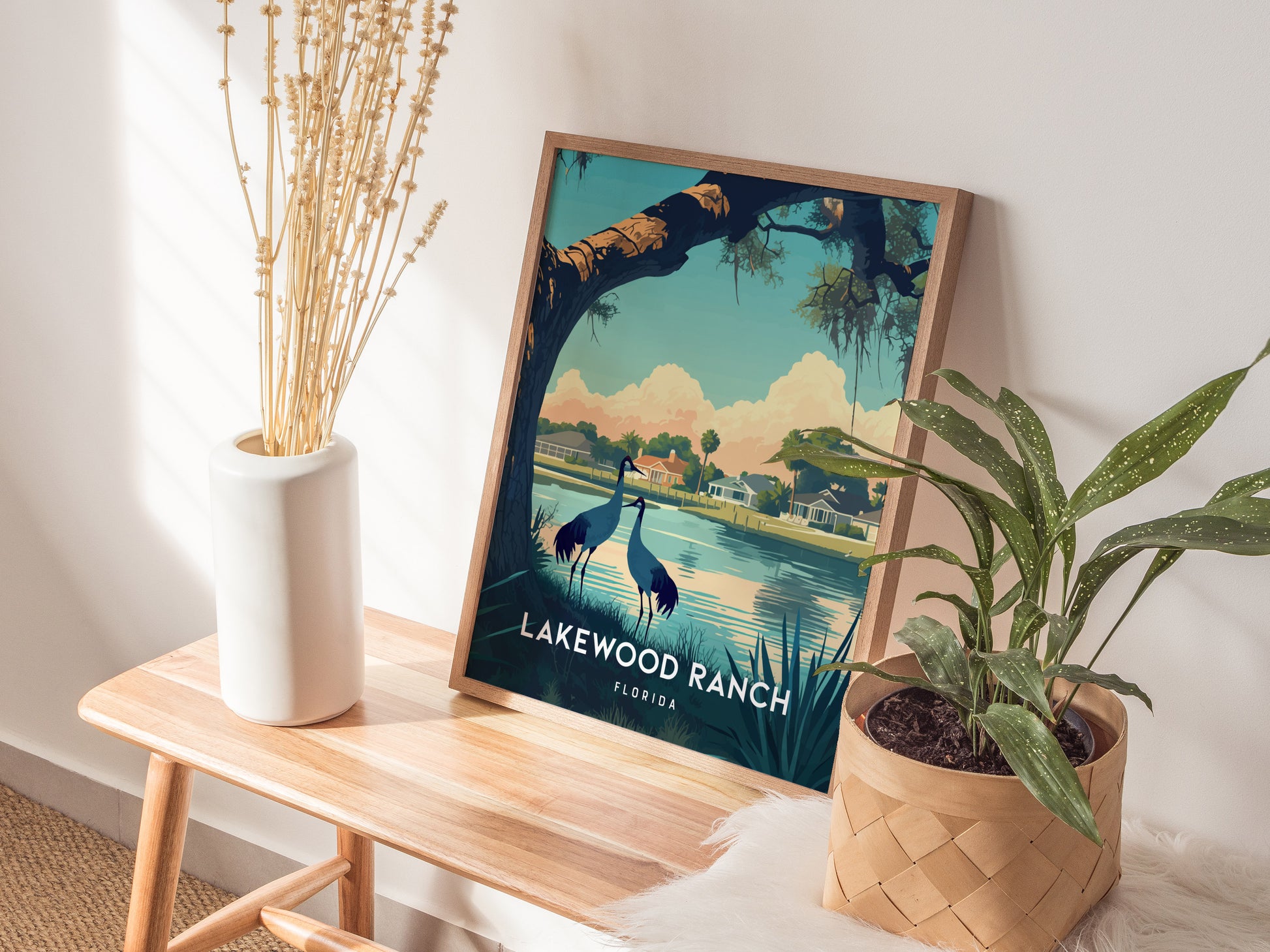 A framed Lakewood Ranch poster featuring a lakeside scene with birds is propped on a wooden bench. Nearby are a white vase with dried stems and a potted plant.