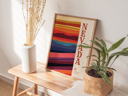 A colorful Nevada state poster with bold stripes leans against a wall on a wooden bench, accompanied by a white vase with dried flowers and a potted plant.