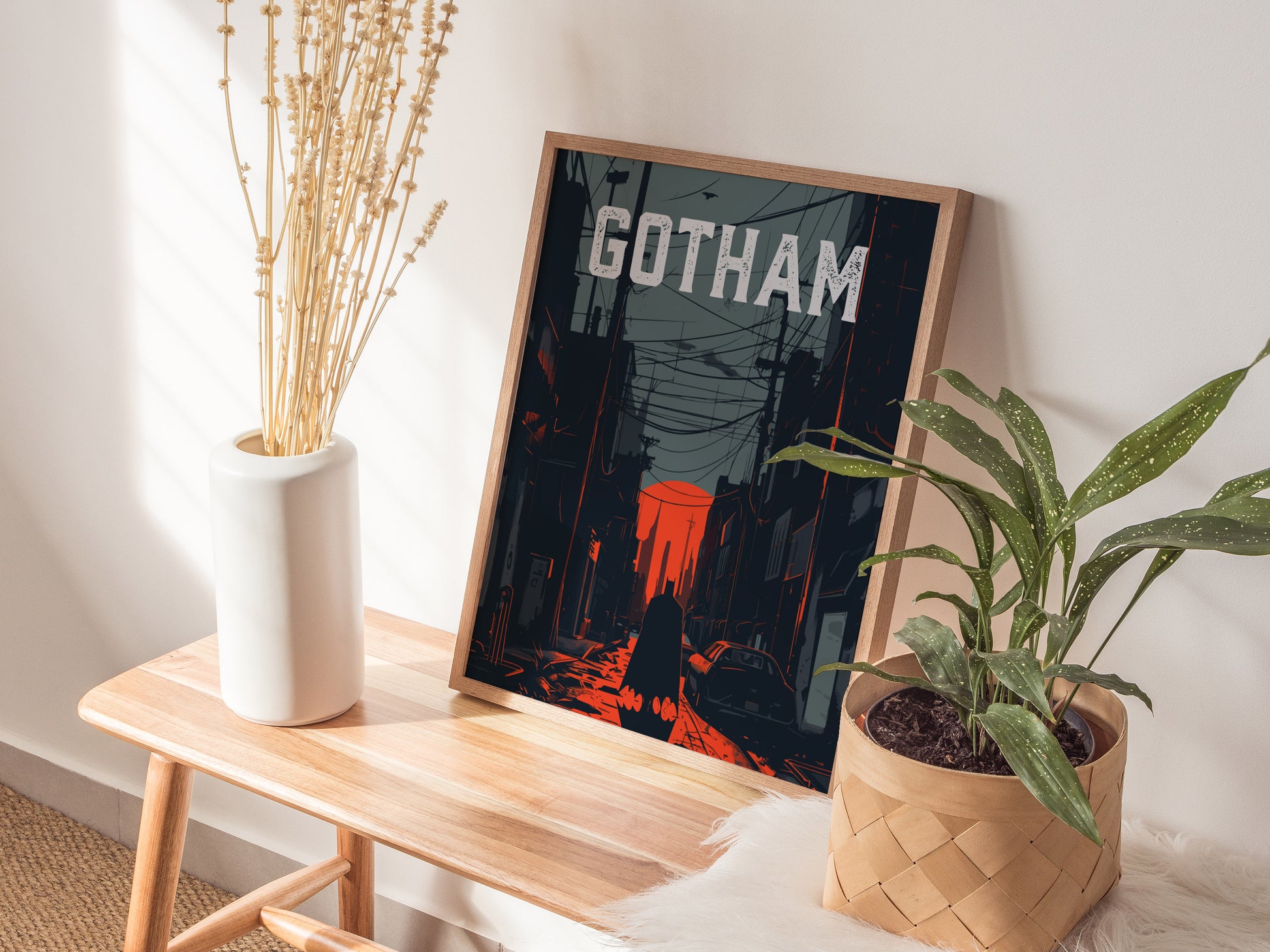 A framed poster titled Gotham is displayed on a wooden bench, featuring a noir-style cityscape in dark tones and red highlights. Beside it is a white vase with tall, dried stems and a green potted plant.