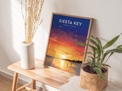 A framed Siesta Key sunset poster with vibrant colors sits on a wooden table beside a white vase with dried flowers and a potted green plant.