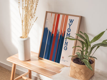 A framed Wyoming poster with red, white, and blue vertical stripes leans against a wall on a wooden bench. Nearby are a white vase with dried stems and a potted.