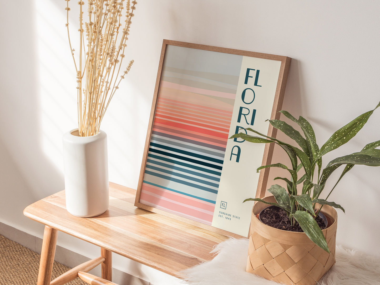 A framed Florida poster with colorful horizontal stripes leans against a wall on a wooden bench, accompanied by a white vase with dried plants and a potted green plant.