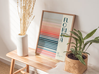 A framed Florida poster with colorful horizontal stripes leans against a wall on a wooden bench, accompanied by a white vase with dried plants and a potted green plant.
