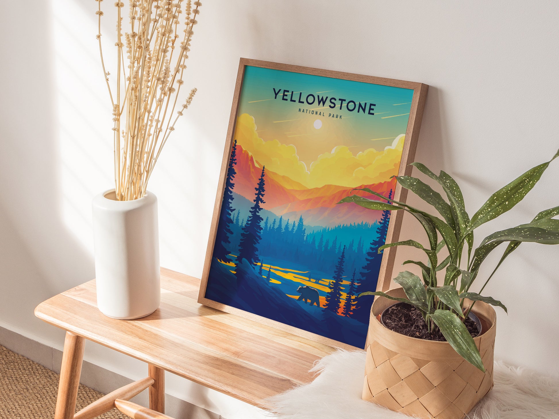 A framed Yellowstone National Park poster with a vibrant sunset and forest scene is displayed on a wooden bench, alongside a white vase with dried stems and a potted plant.