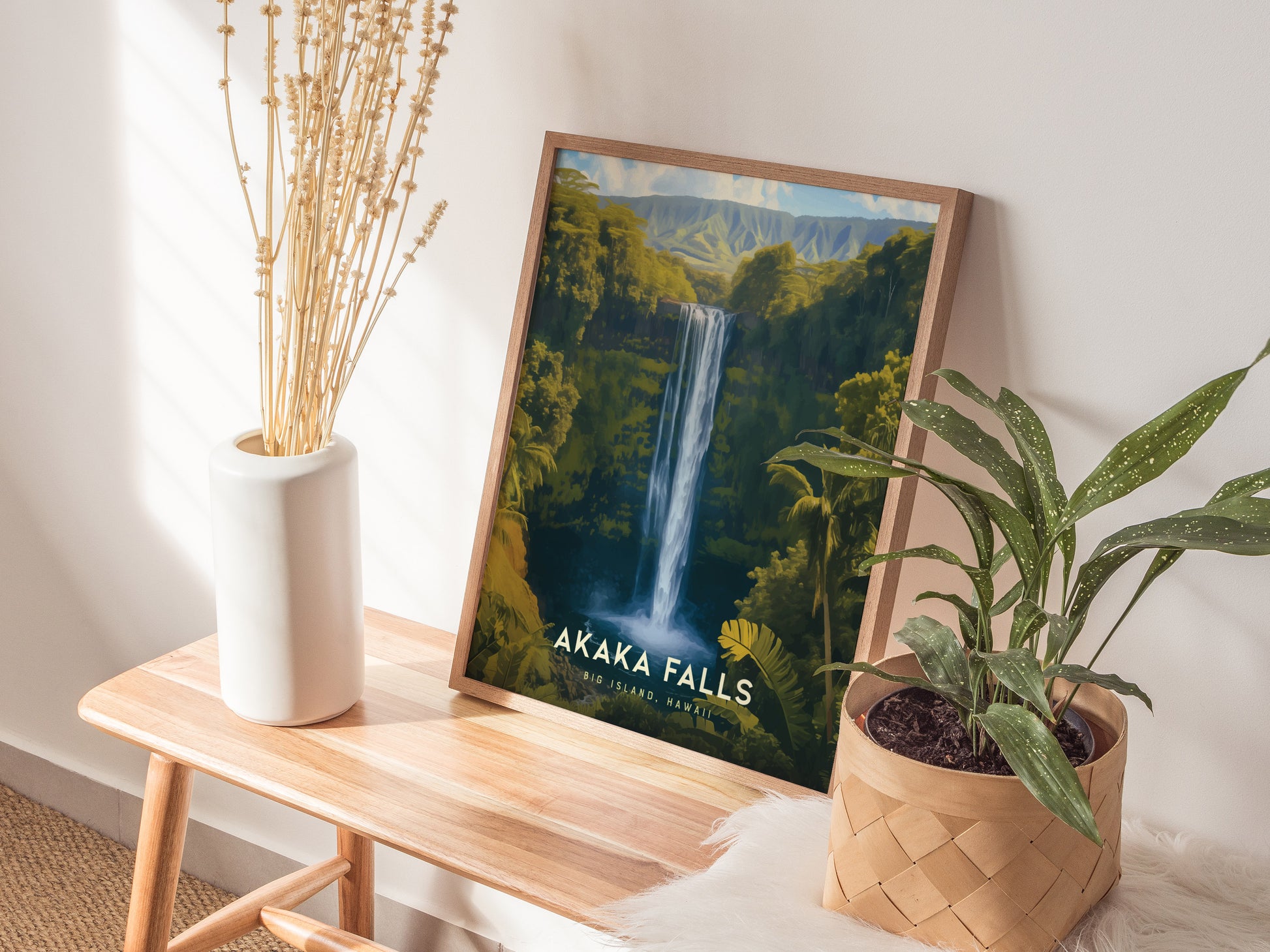 A framed poster of Akaka Falls rests on a wooden bench, accompanied by a white vase with dried flowers and a potted plant. Sunlight casts soft shadows on the wall.