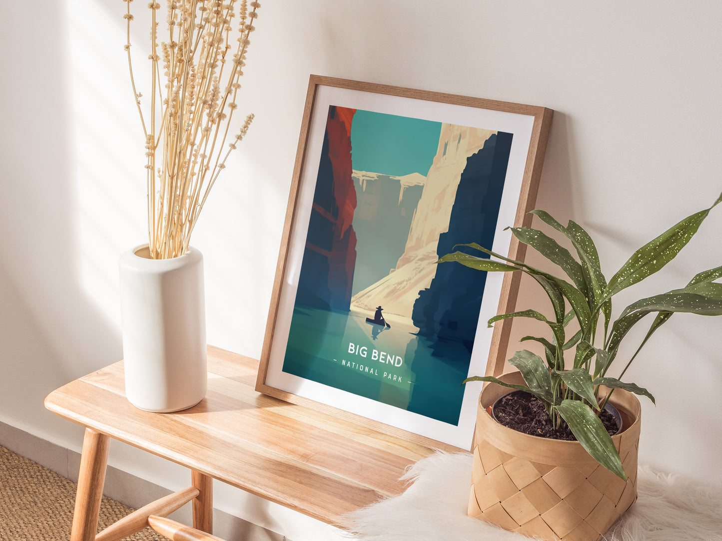 A framed Big Bend National Park poster rests on a wooden bench beside a white vase with dried flowers and a green potted plant, creating a cozy decor setting.