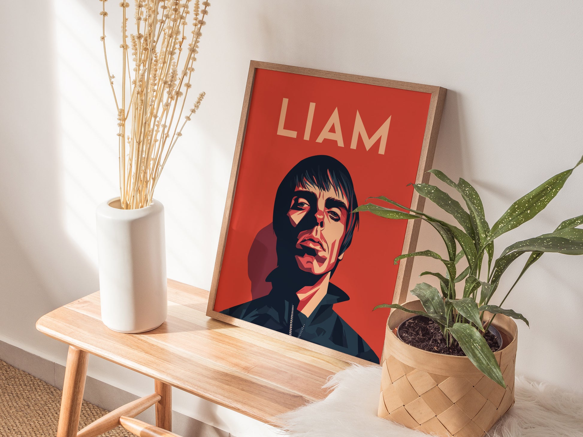 A framed poster with a stylized portrait on a red background labeled LIAM sits on a wooden bench, accompanied by a white vase and a potted plant.