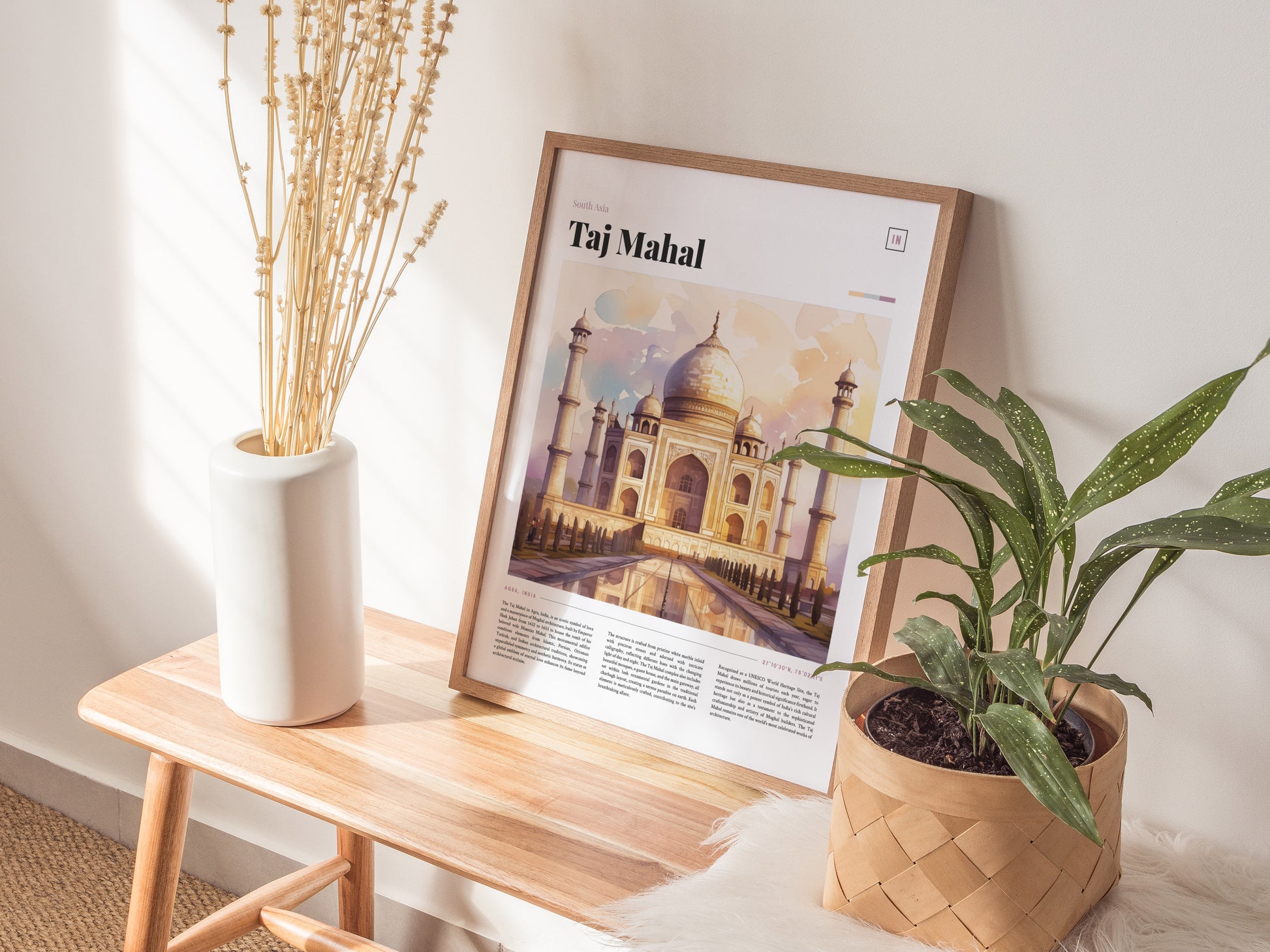 A framed Taj Mahal poster rests on a wooden bench, flanked by a white vase with dried flowers and a potted green plant. Natural light enhances the serene setting.