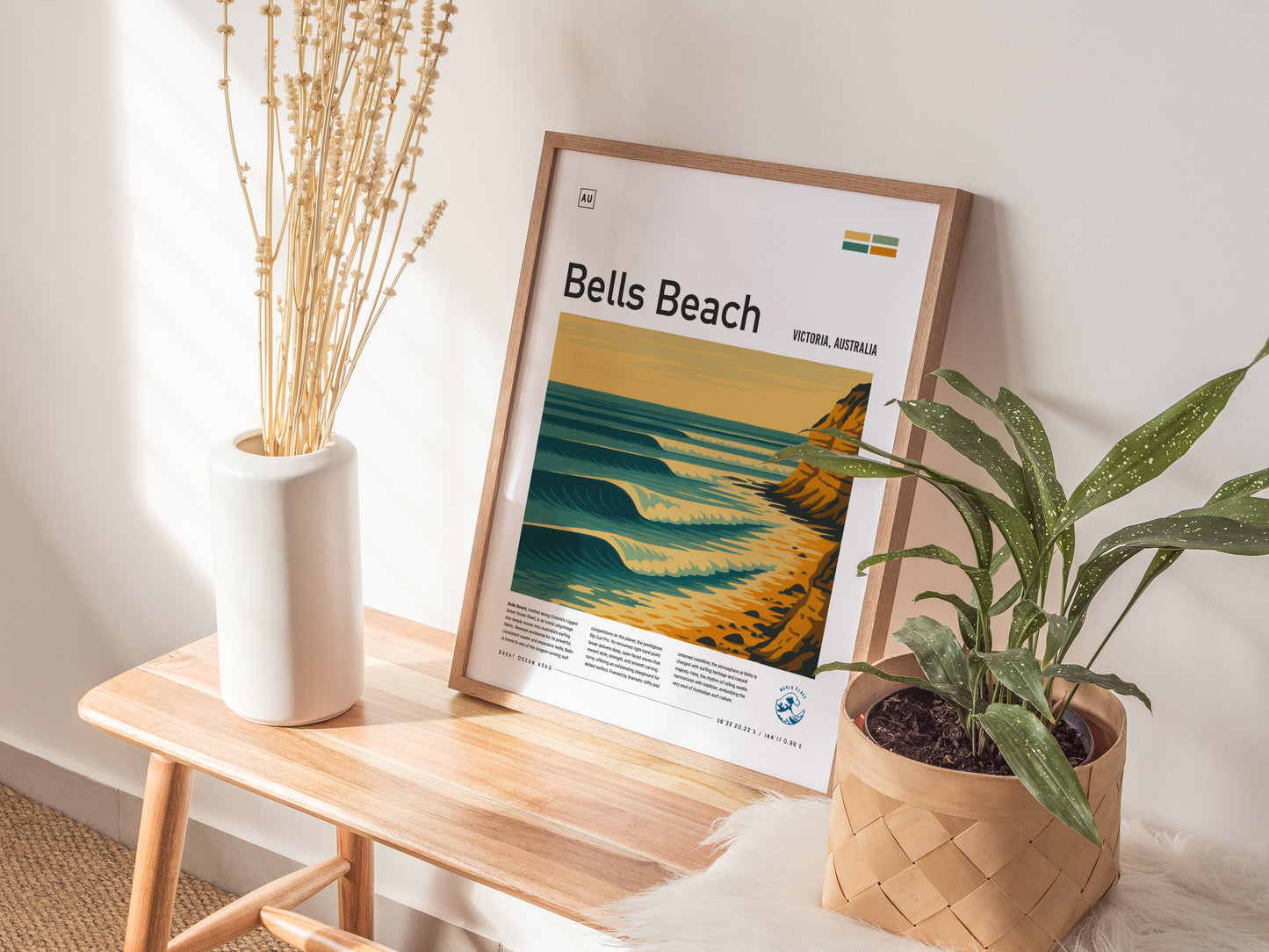 Framed Bells Beach Surf Spot poster leaning against a wall on a wooden table, accompanied by a white vase with dried flowers and a green potted plant.