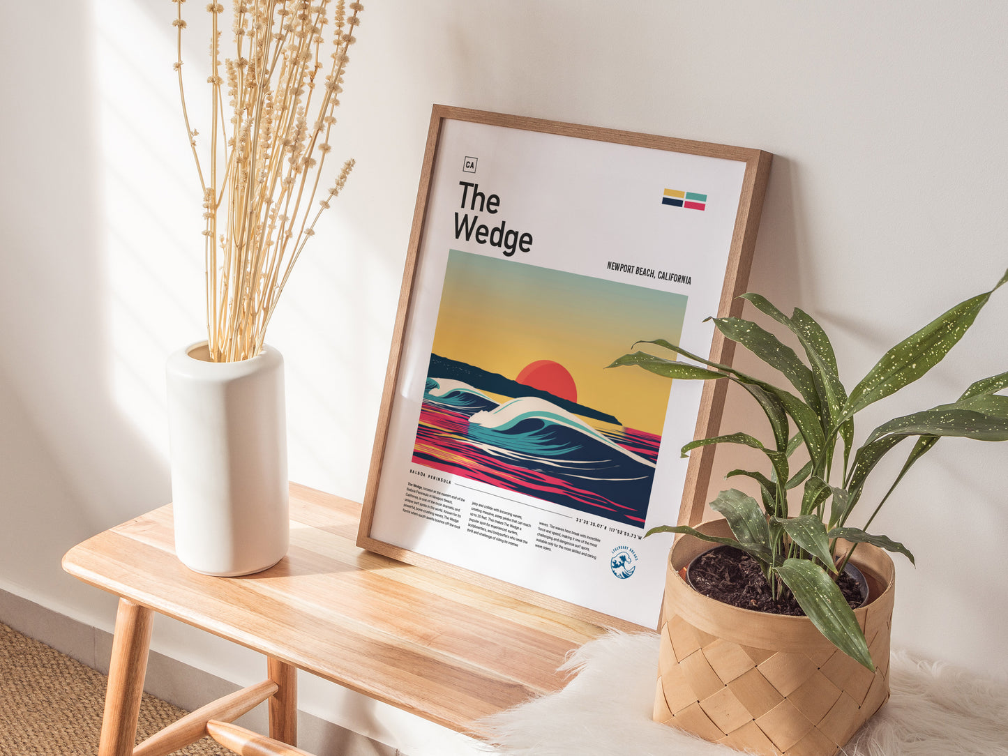 A framed poster titled The Wedge leans against a wall on a wooden bench, flanked by a white vase with dried flowers and a potted plant.