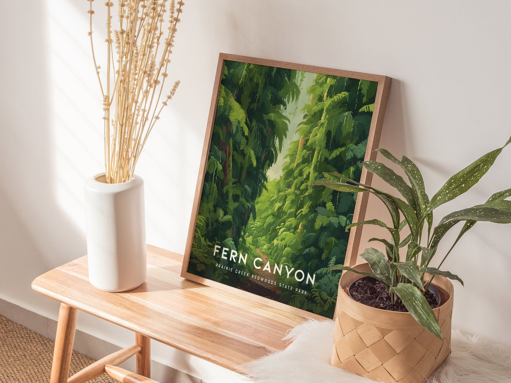 A framed poster titled Fern Canyon rests on a wooden bench, surrounded by a white vase with dried flowers and a potted green plant. Natural light illuminates the scene.
