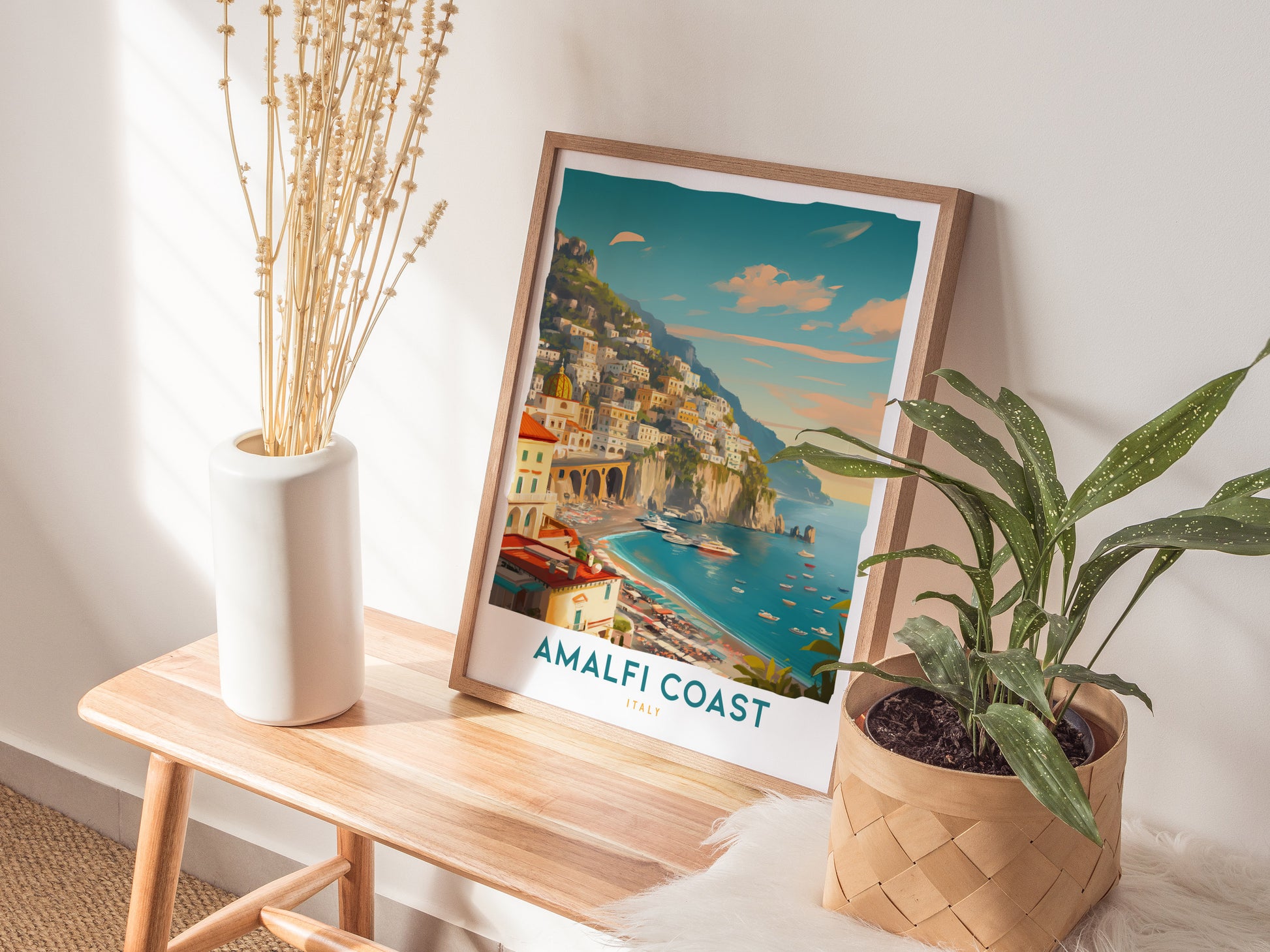 A framed Amalfi Coast travel print rests on a wooden bench beside a white vase with dried stems and a potted plant, bathed in warm sunlight.
