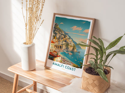 A framed Amalfi Coast travel print rests on a wooden bench beside a white vase with dried stems and a potted plant, bathed in warm sunlight.