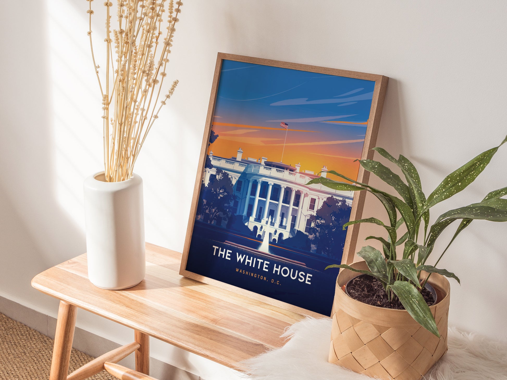 A framed poster of the White House in vibrant colors sits on a wooden bench, surrounded by a white vase with dried flowers and a potted green plant.