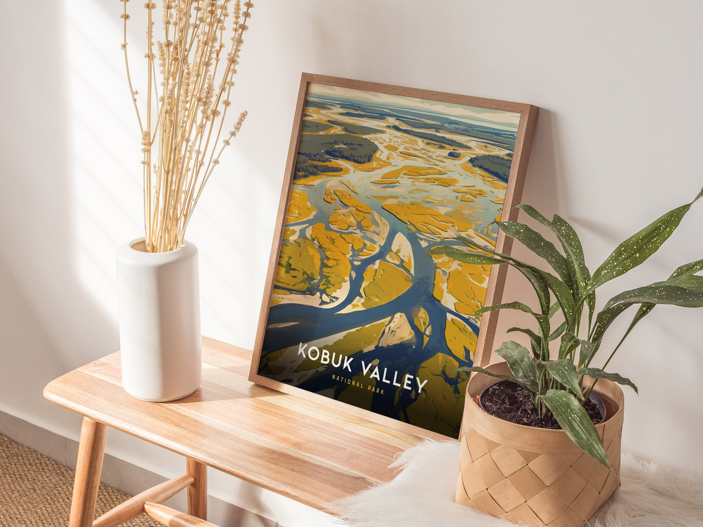 A framed poster of Kobuk Valley rests on a wooden bench, accompanied by a white vase with dried plants and a potted green plant, creating a cozy.