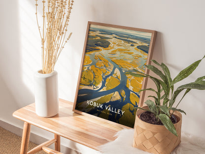 A framed poster of Kobuk Valley rests on a wooden bench, accompanied by a white vase with dried plants and a potted green plant, creating a cozy.