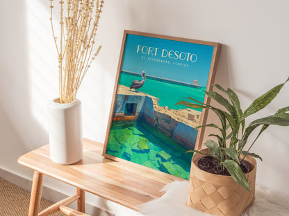 The image shows a framed Fort Desoto poster on a wooden bench, accompanied by a white vase with dried flowers and a potted green plant. Sunlight casts soft shadows.
