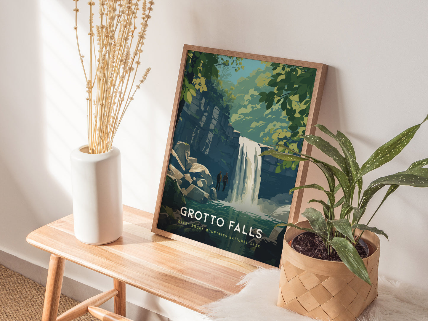 A framed Grotto Falls poster is displayed on a wooden bench beside a white vase with dried flowers and a potted green plant. The poster features a waterfall scene with lush greenery, creating a serene and natural setting.