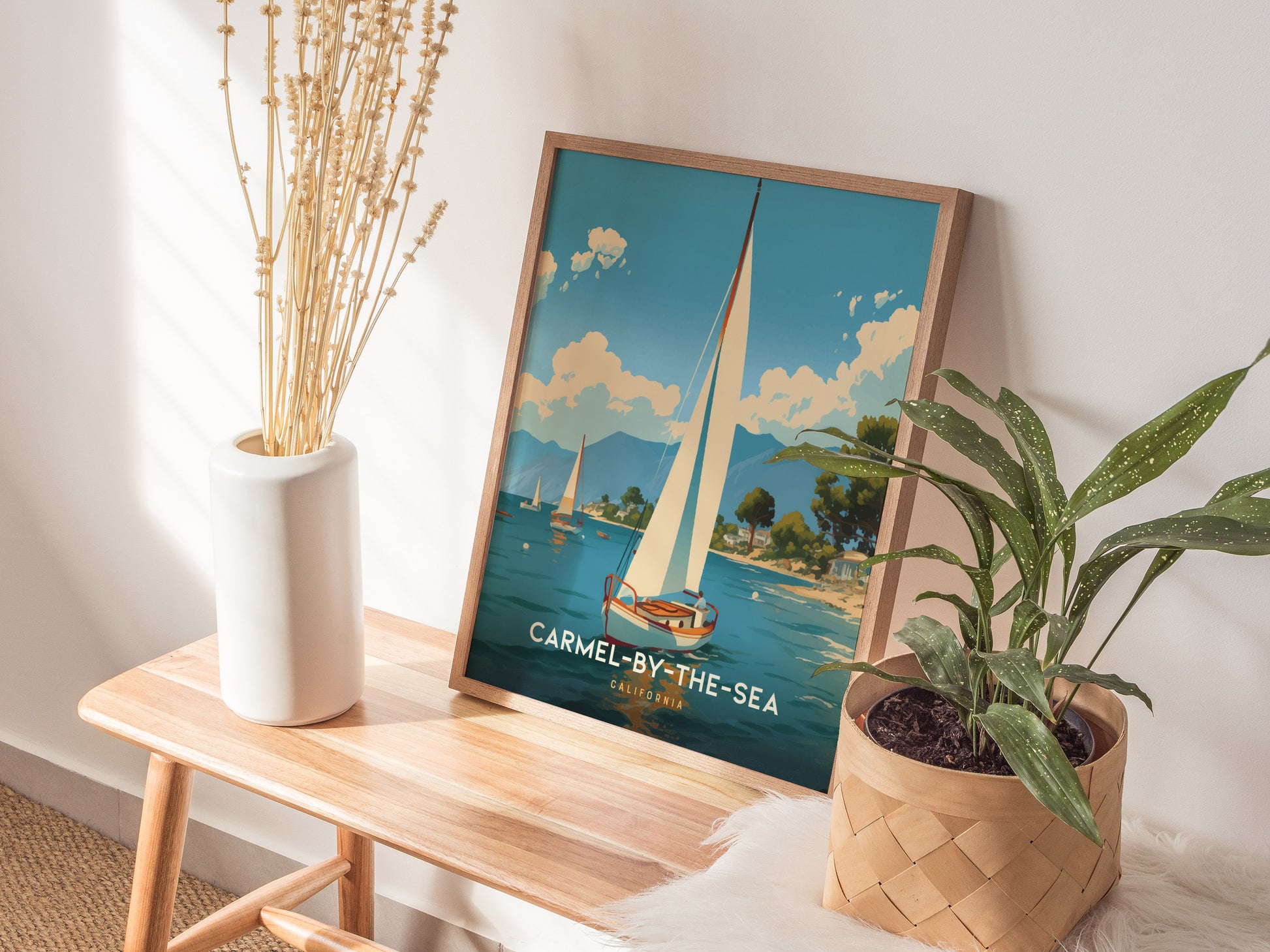A framed poster titled Carmel-by-the-Sea featuring a sailboat is displayed on a wooden bench, accompanied by a white vase with dried flowers and a potted plant.