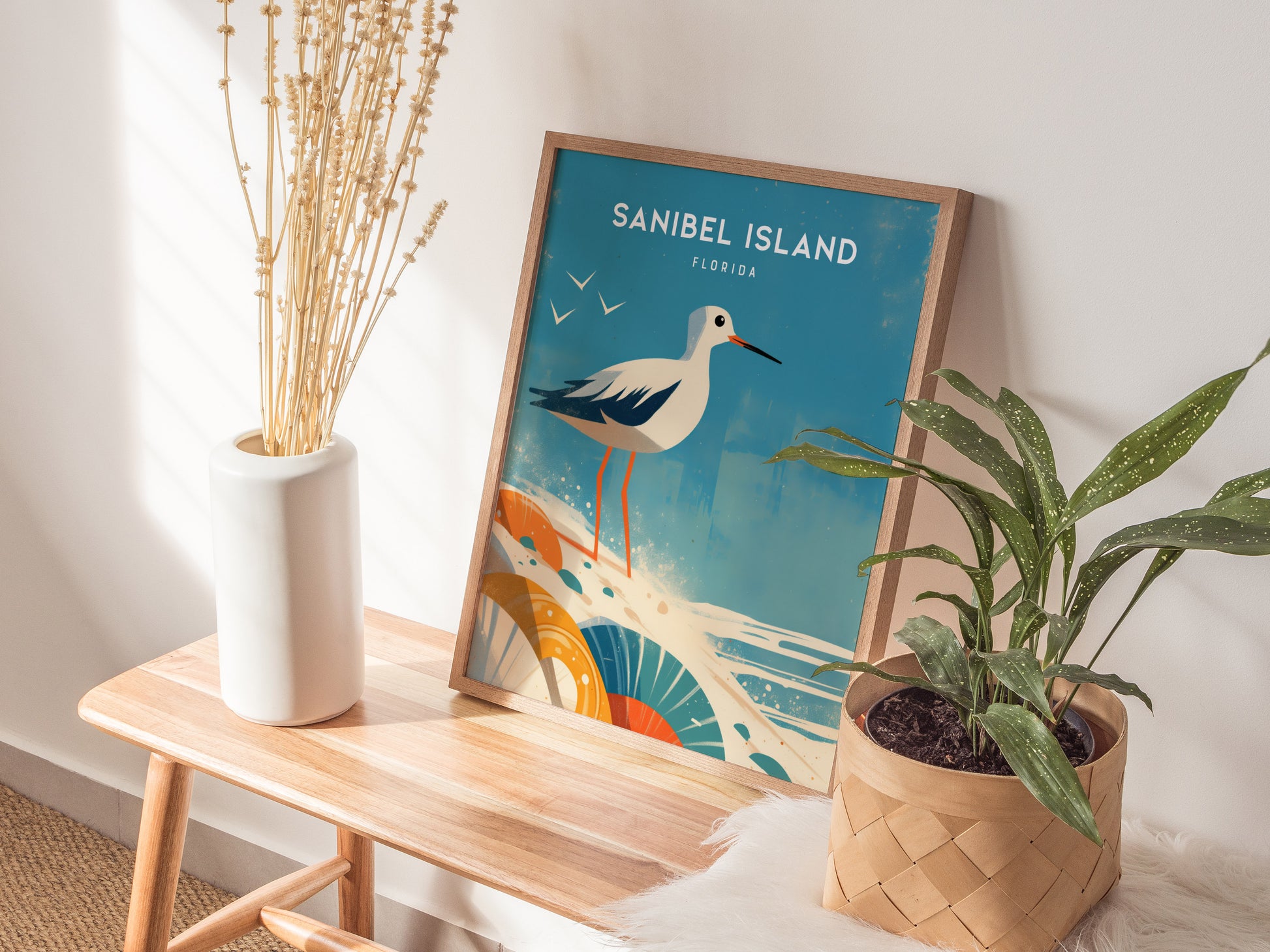 A Sanibel Island poster featuring a sandpiper and seashells is displayed on a wooden bench, alongside a white vase with dried flowers and a potted green plant.