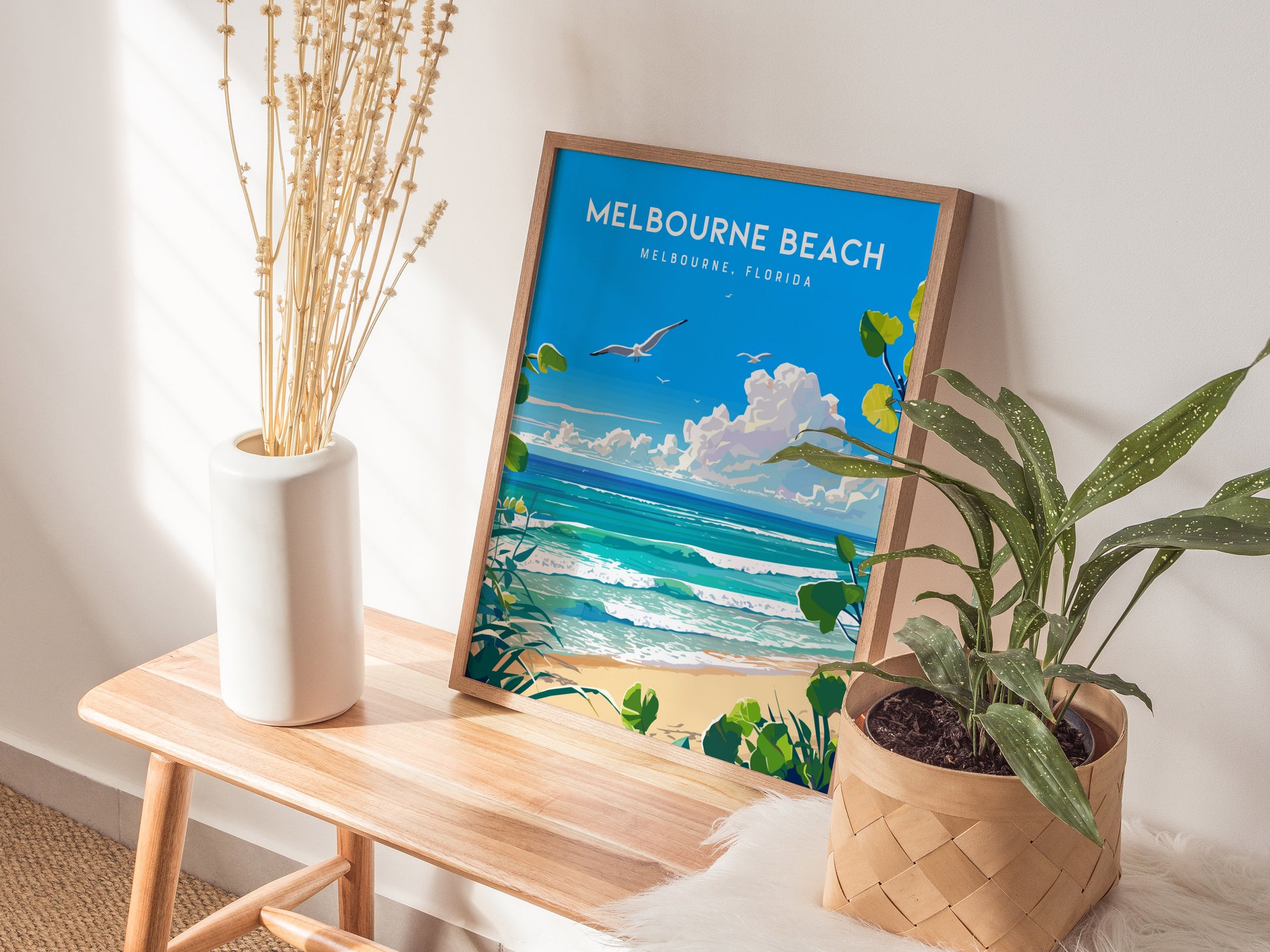 A framed Melbourne Beach poster depicting a serene ocean scene rests on a wooden bench, accompanied by a white vase of dried flowers and a potted green plant.