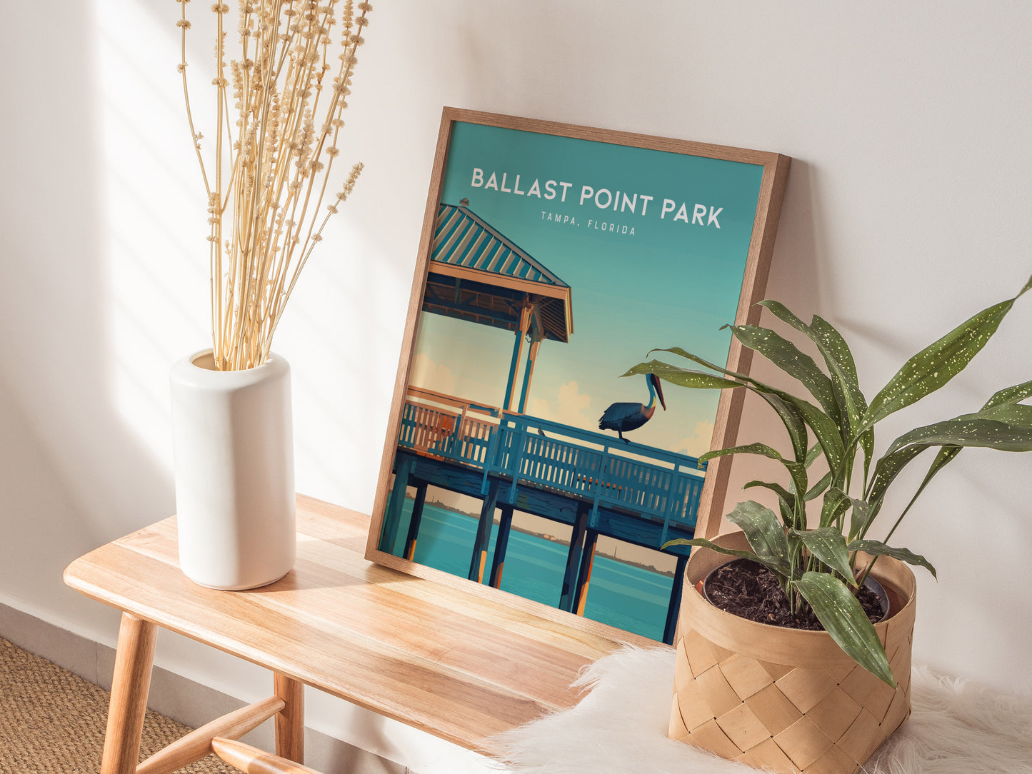 A framed poster of Ballast Point Park in Tampa, Florida, featuring a pelican on a pier, is displayed on a wooden table with a white vase and green plant.