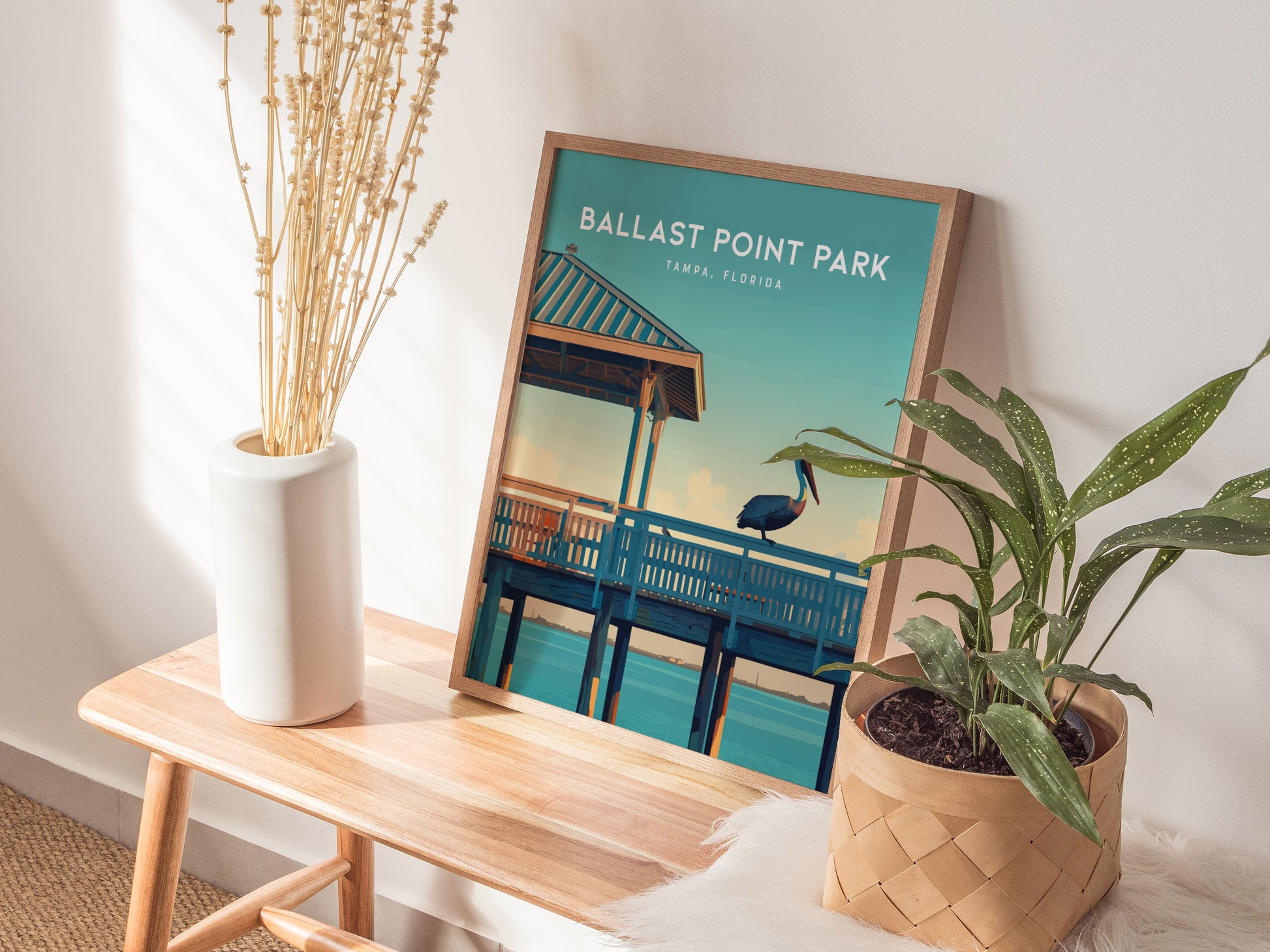 A framed poster of Ballast Point Park in Tampa, Florida, featuring a pelican on a pier, is displayed on a wooden table with a white vase and green plant.