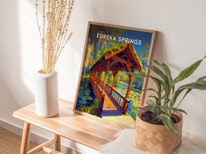 A framed Eureka Springs poster showing a wooden bridge over a stream is displayed on a wooden bench, next to a white vase with dried flowers and a potted plant.