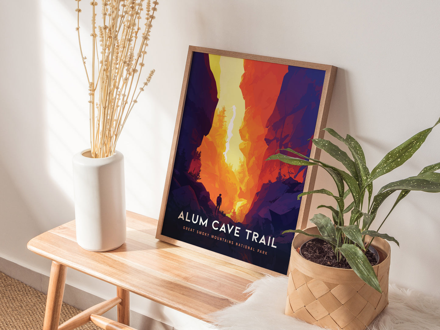 A framed Alum Cave Trail poster with vibrant, fiery colors leans against a wall on a wooden bench. A white vase with dried flowers and a potted plant are nearby.