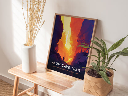 A framed Alum Cave Trail poster with vibrant, fiery colors leans against a wall on a wooden bench. A white vase with dried flowers and a potted plant are nearby.