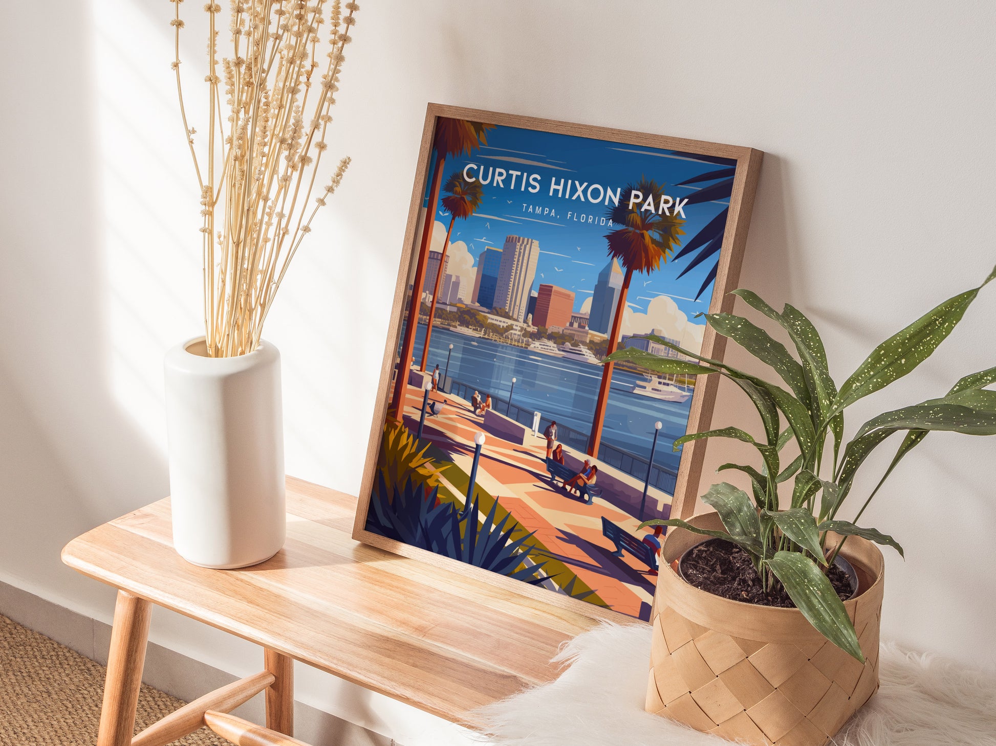 A Curtis Hixon Park poster rests on a wooden table, surrounded by a white vase with dried flowers and a potted plant, creating a cozy interior scene.