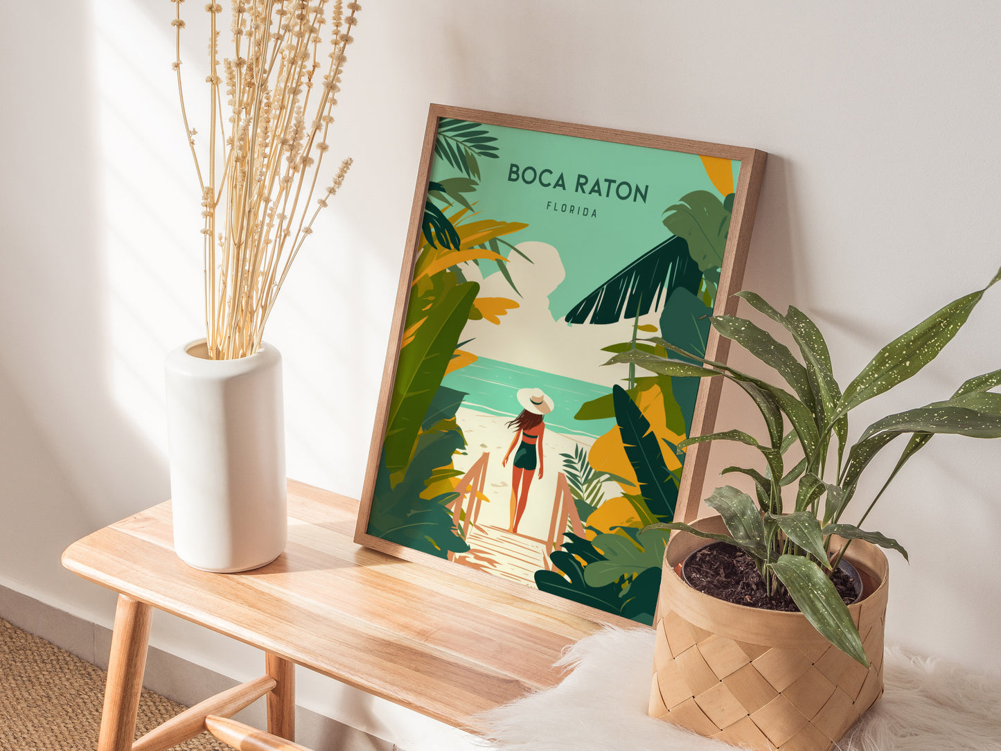 A framed Boca Raton travel print is displayed on a wooden bench next to a white vase with dried flowers and a potted plant. Soft sunlight illuminates the scene.