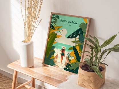 A framed Boca Raton travel print is displayed on a wooden bench next to a white vase with dried flowers and a potted plant. Soft sunlight illuminates the scene.