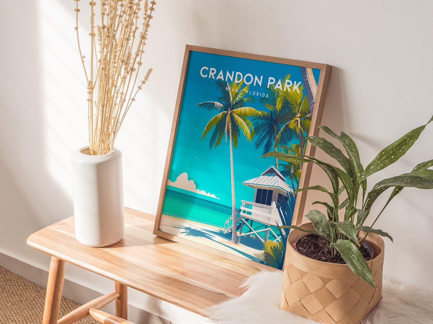 A framed Crandon Park Beach poster rests on a wooden bench, featuring palm trees and a lifeguard tower against a vibrant blue sky. Beside it, a white vase with dried stems and a potted plant add a touch of nature.