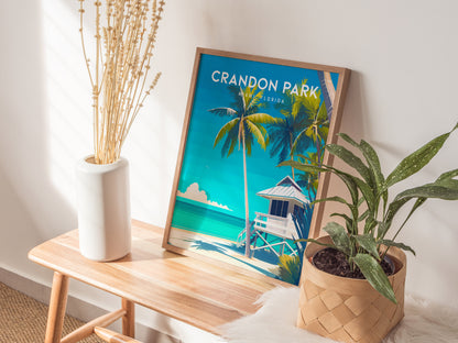 A framed Crandon Park Beach poster rests on a wooden bench, featuring palm trees and a lifeguard tower against a vibrant blue sky. Beside it, a white vase with dried stems and a potted plant add a touch of nature.