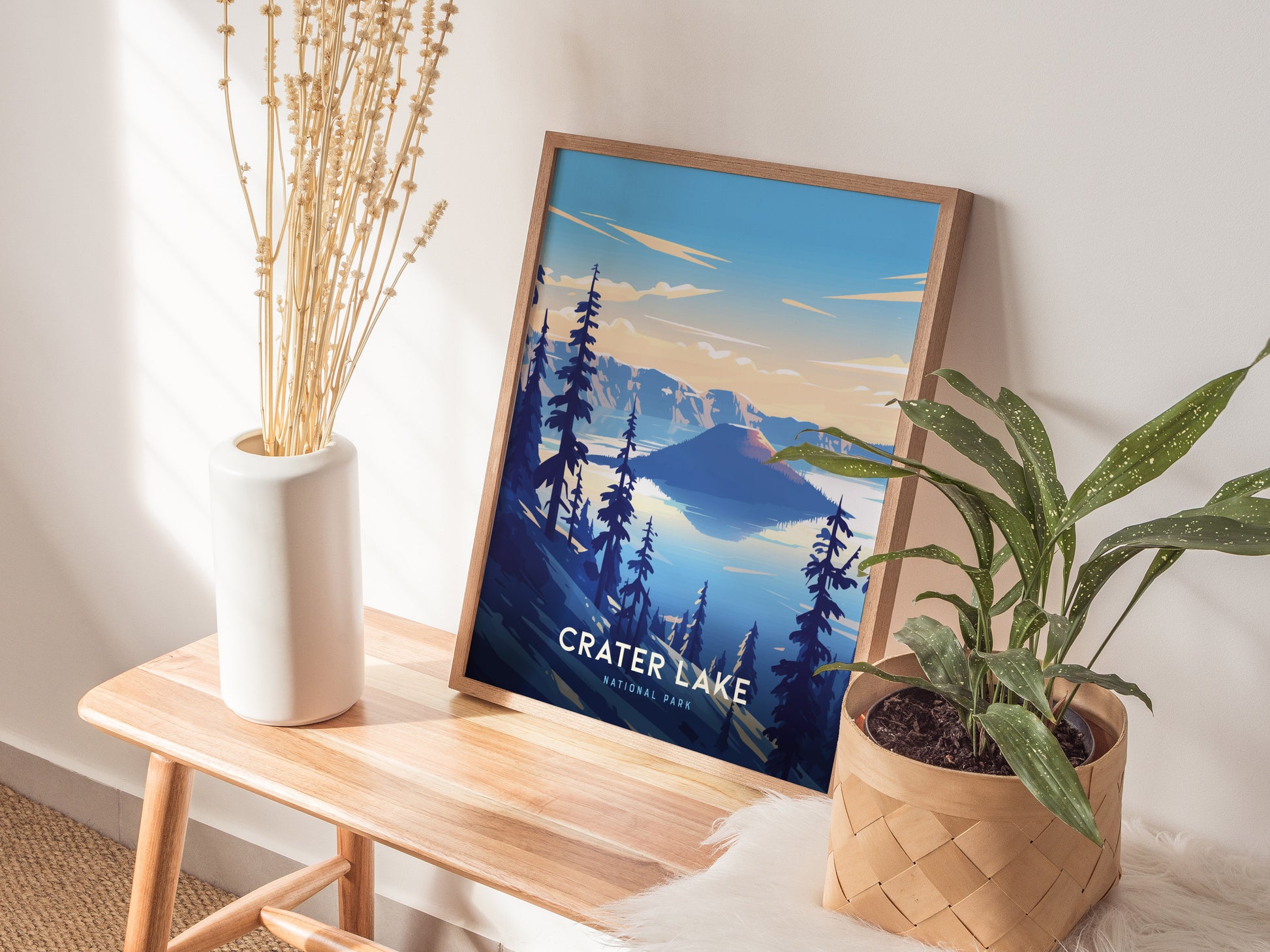 A framed Crater Lake National Park poster sits on a wooden bench, flanked by a tall white vase with dried stems and a potted green plant.