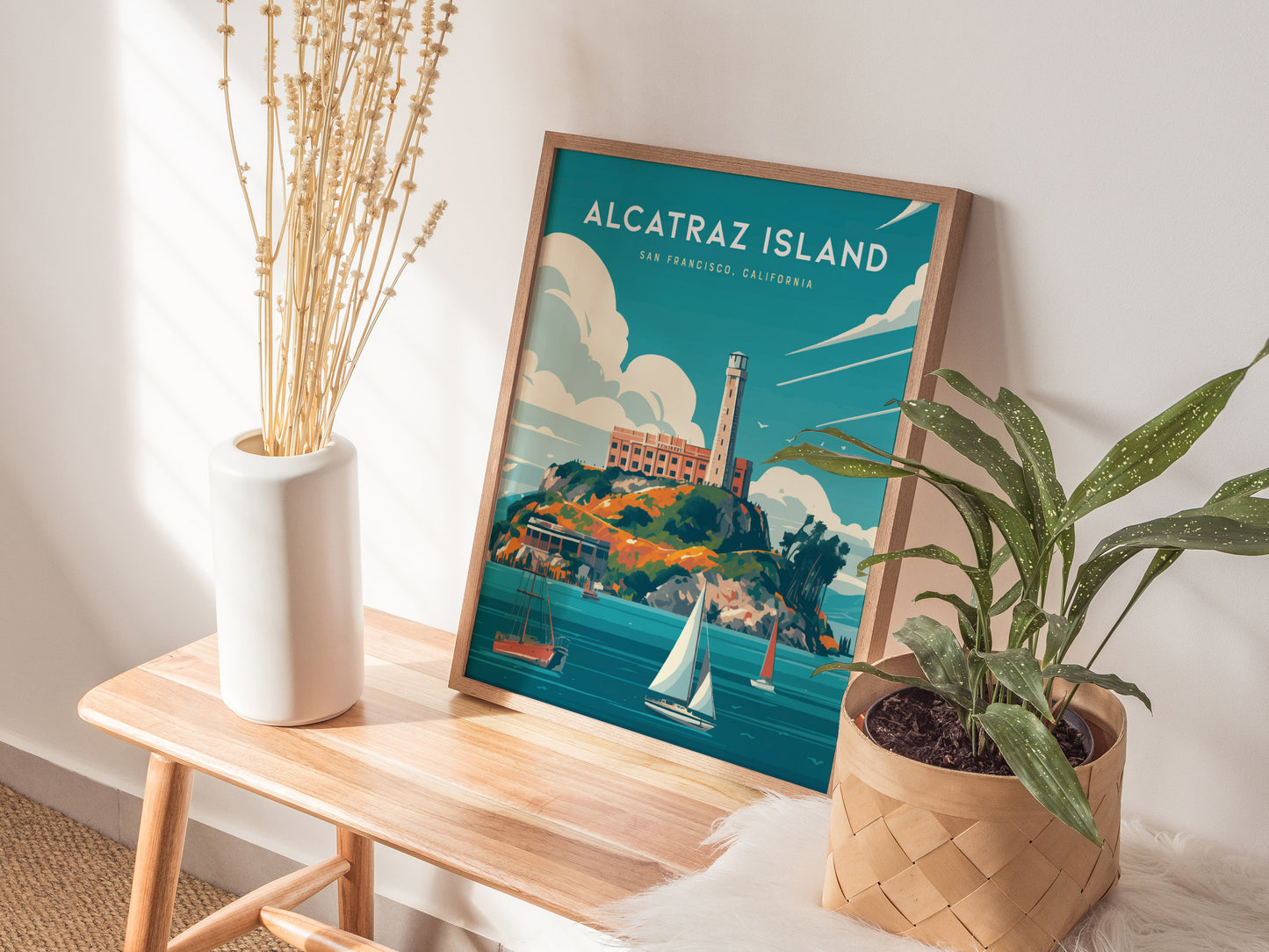 A framed Alcatraz Island poster sits on a wooden bench next to a white vase with dried flowers and a potted green plant.