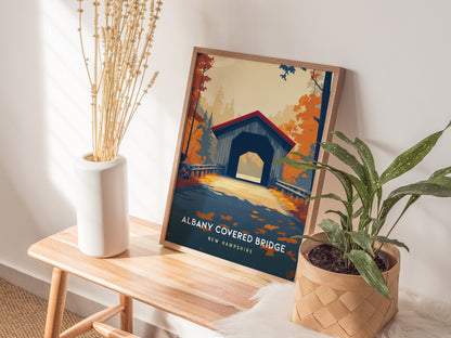 A framed poster of the Albany Covered Bridge in New Hampshire is displayed on a wooden table in a bright room.
