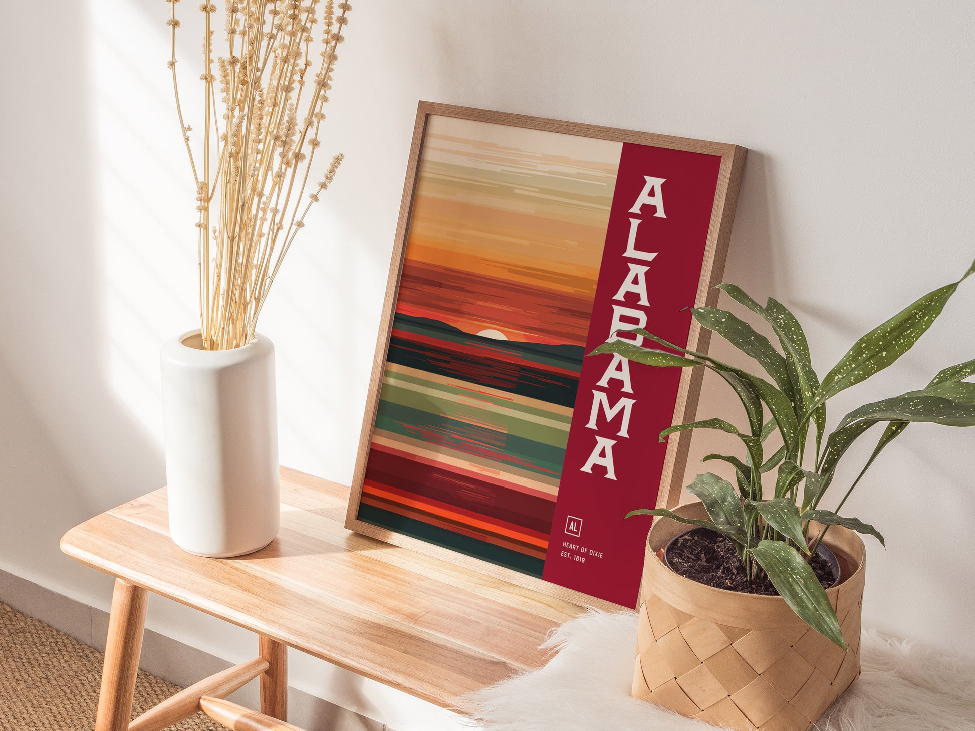 A minimalist Alabama-themed poster with abstract warm hues is displayed on a wooden bench, accompanied by a white vase with dried flowers and a potted plant.